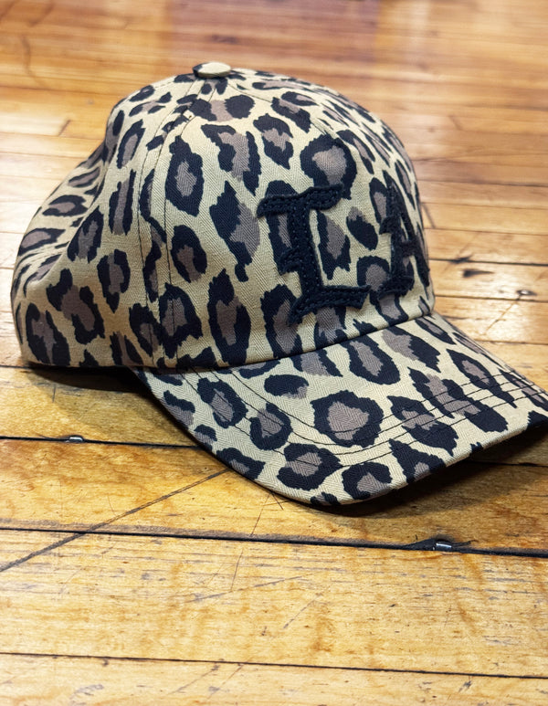 A leopard print baseball cap with a black embroidered letter 'I' on the front, resting on a wooden plank surface.