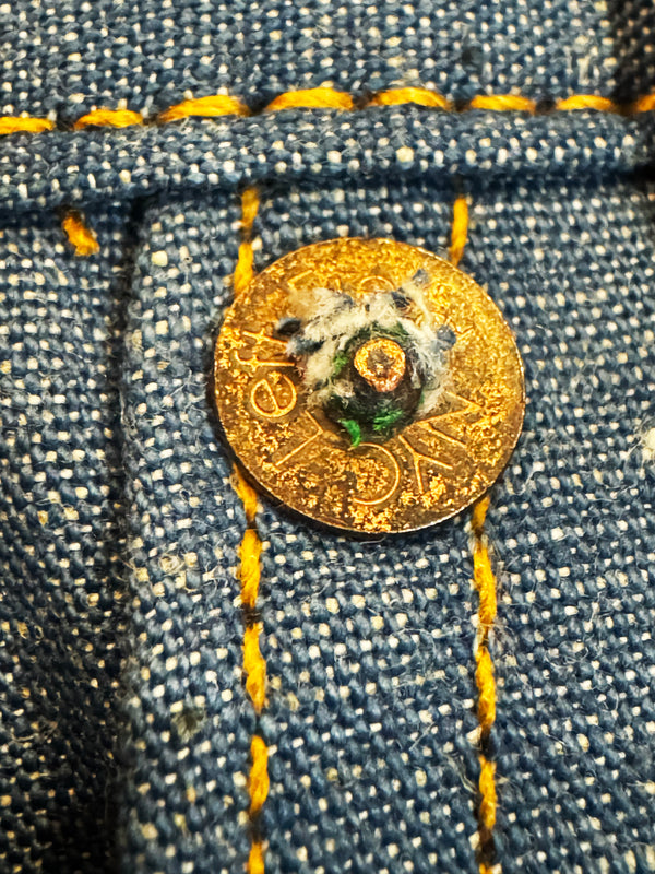 A close-up of a gold-toned button with a decorative floral center, sewn onto blue denim fabric with yellow contrast stitching.
